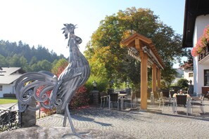 A courtyard with a metal rooster sculpture, outdoor seating, and a wooden pergola. - Landhotel und Gasthof Schmalhofer (Tittling)
