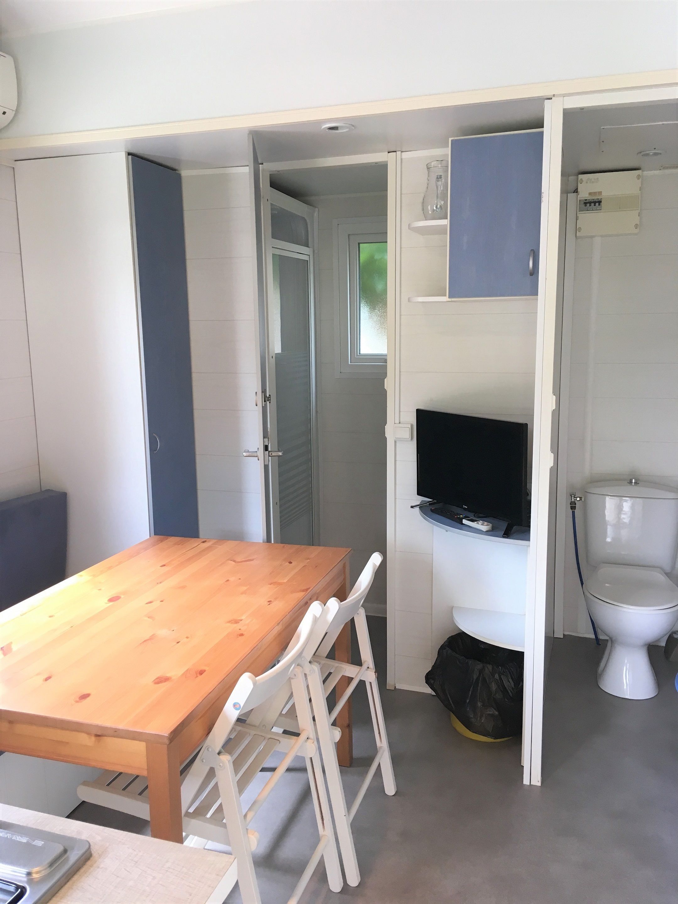 A compact room with a wooden table and white chairs, a television, and a toilet.