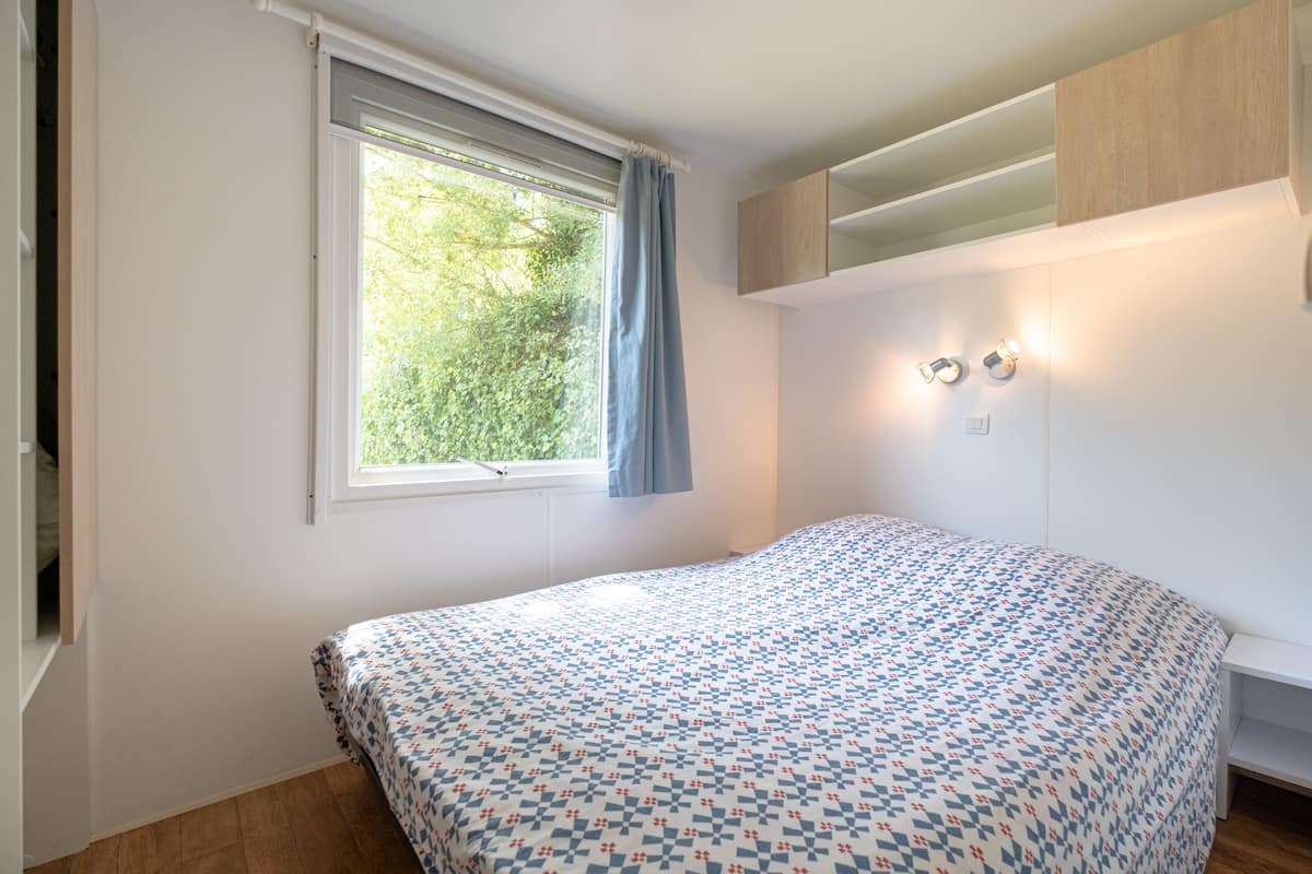 Un dormitorio con una cama, una ventana con cortinas, una lámpara de pared y un estante empotrado.