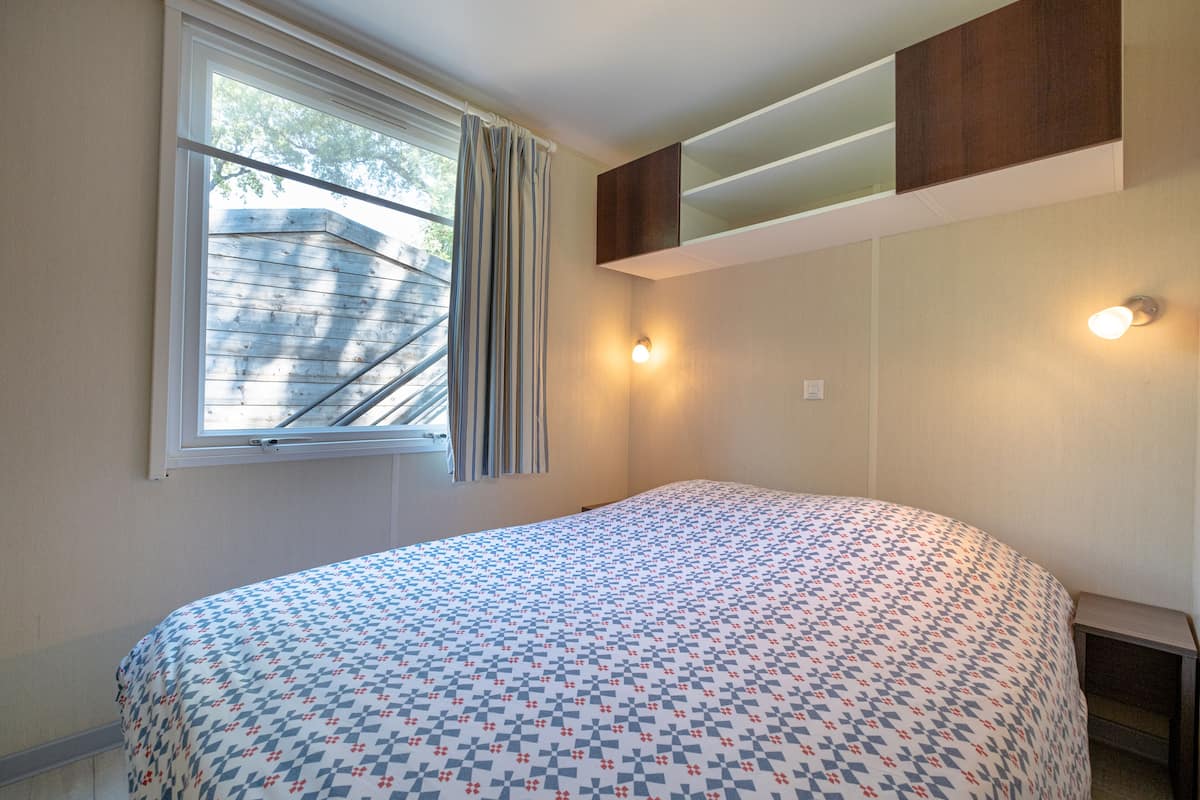 Un dormitorio con cama, una ventana con cortinas, un estante montado en la pared y dos apliques.