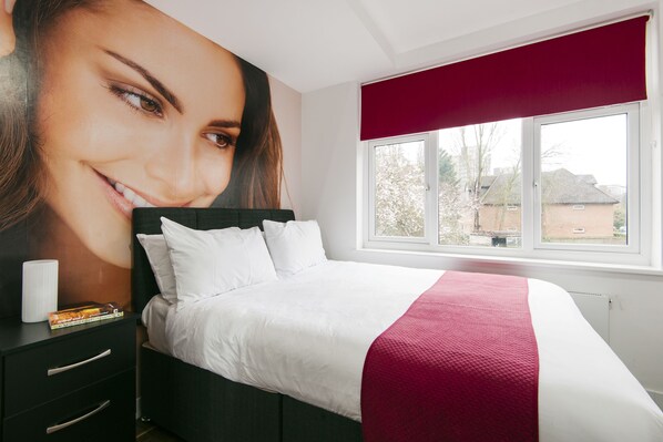 A bedroom with a large bed, a nightstand with books, a window with a red blind, and a view of houses outside.
