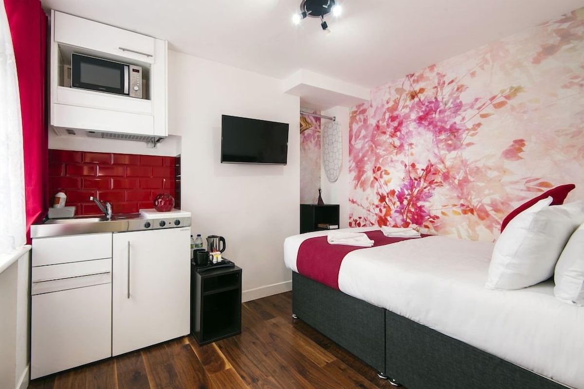 A compact hotel room with a kitchenette, a bed with a floral wall mural, and a flat-screen TV.
