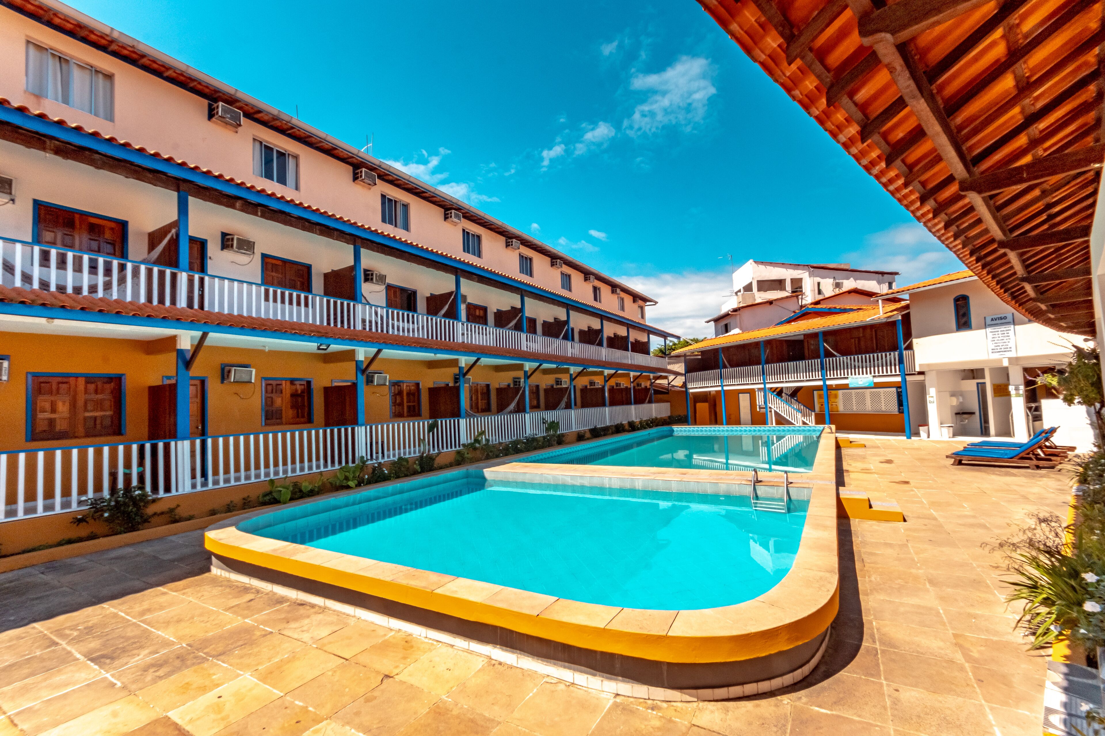 A swimming pool with a tiled deck, a covered area with a roof, and a building with balconies and windows.