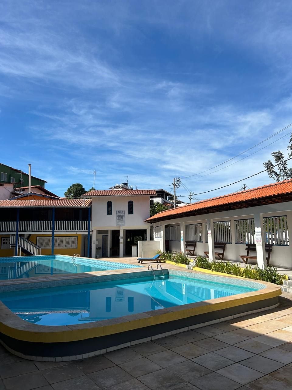 Une piscine entourée d’une terrasse carrelée, elle-même bordée de bâtiments aux toits recouverts de tuiles rouges, le tout sous un ciel bleu azur.