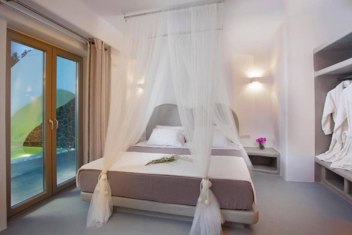 Un dormitorio con cama, mesita de noche, ventana con cortinas y vistas a un paisaje verde.