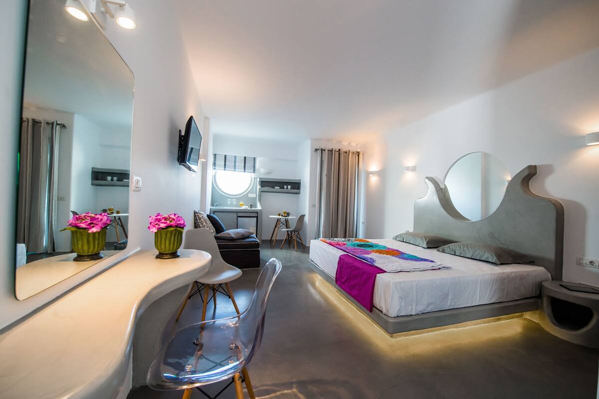 Habitación de hotel moderna con una cama grande, un escritorio, una silla, un televisor y un baño visible a través de un panel de vidrio.