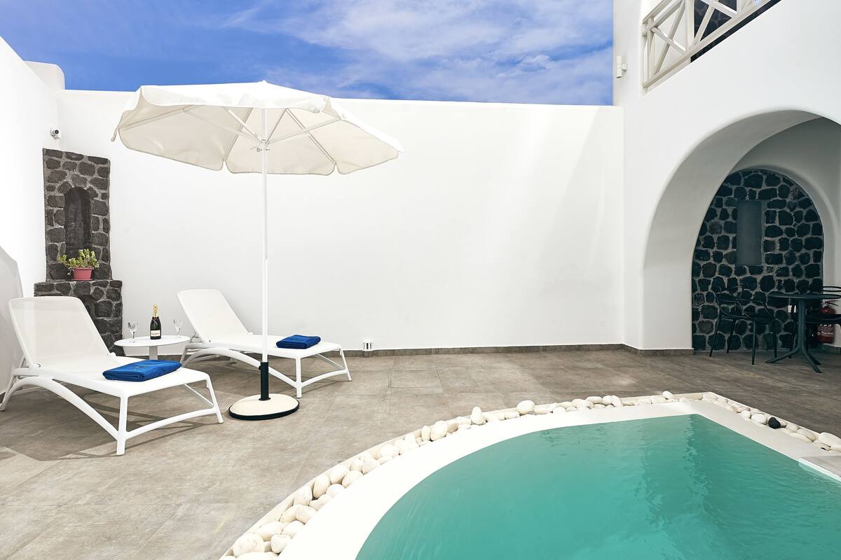 Un área junto a la piscina con sillones blancos, una sombrilla blanca y un muro de piedra.