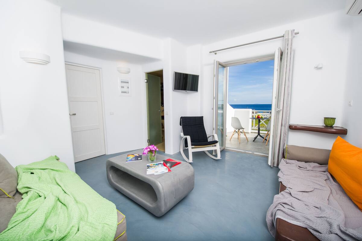 Una sala de estar moderna con un sofá gris, una manta verde, una silla blanca y un balcón con vista al mar.