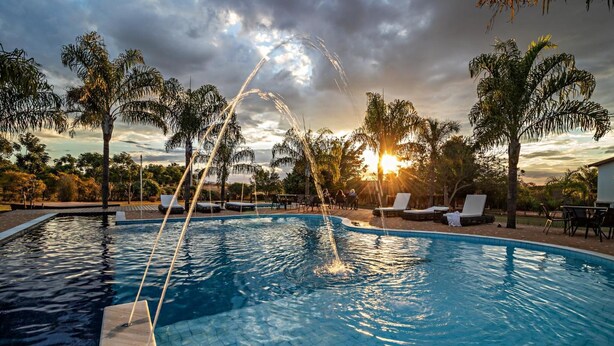A swimming pool with water jets, surrounded by palm trees and lounge chairs, with a sunset in the background. - Pousada Pé na Terra (Brotas)