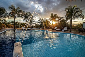 A swimming pool with water jets, surrounded by palm trees and lounge chairs, with a sunset in the background. - Pousada Pé na Terra (Brotas)