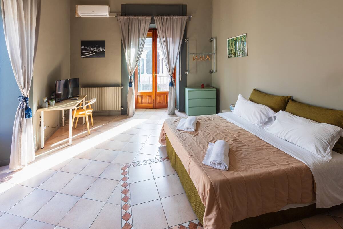 Habitación de hotel con una cama, un escritorio con silla, un televisor, una mesita de noche verde y una ventana con cortinas.