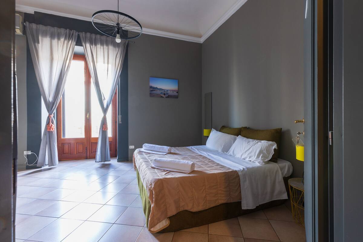 Una habitación de hotel con una cama, cortinas, un cuadro en la pared y una puerta.