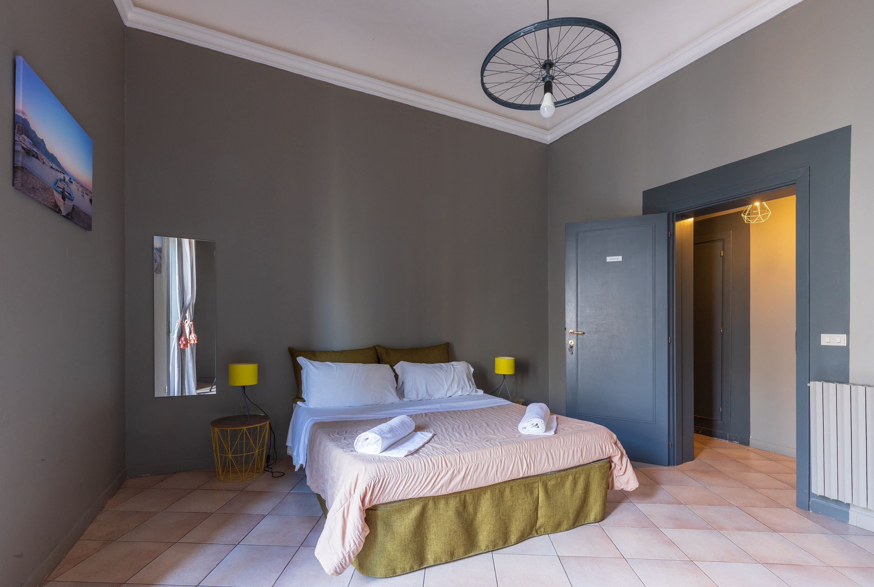 A bedroom with a bed, a mirror, a lamp, and a door leading to another room.