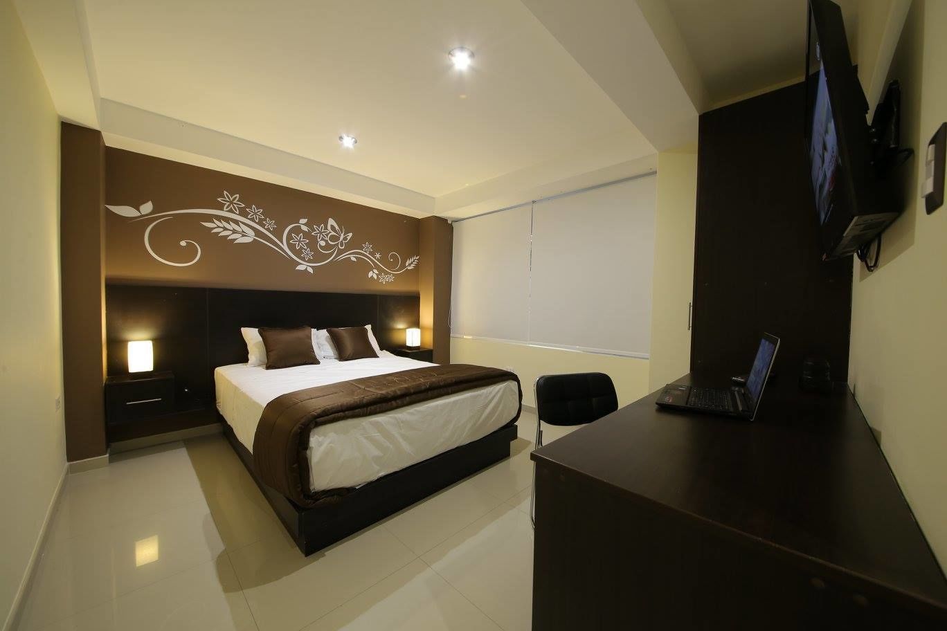 A modern hotel room with a large bed, a desk with a laptop, a flat-screen TV, and a decorative wall design.