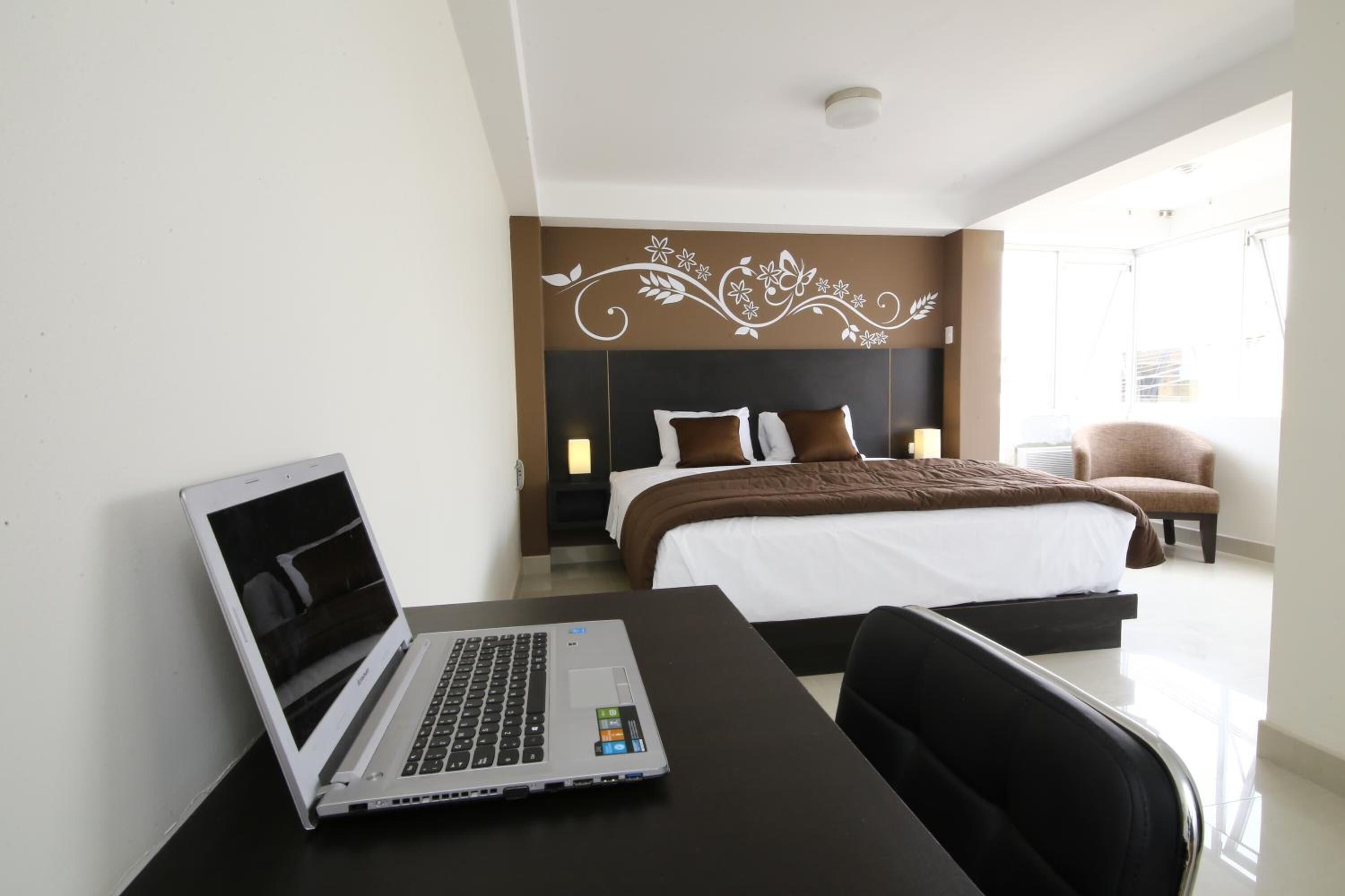 A hotel room with a bed, a desk with a laptop, a chair, and a decorative wall.