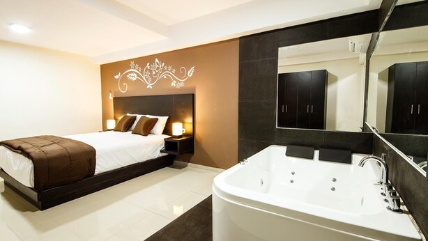 Un dormitorio moderno con una cama grande, una mesita de noche con lámpara, un espejo amplio y un baño separado con bañera y muebles de tonos oscuros.