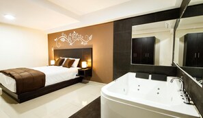 Quarto moderno com uma cama grande, mesa de cabeceira com abajur, espelho amplo e banheiro separado com banheira e armários em tons escuros.