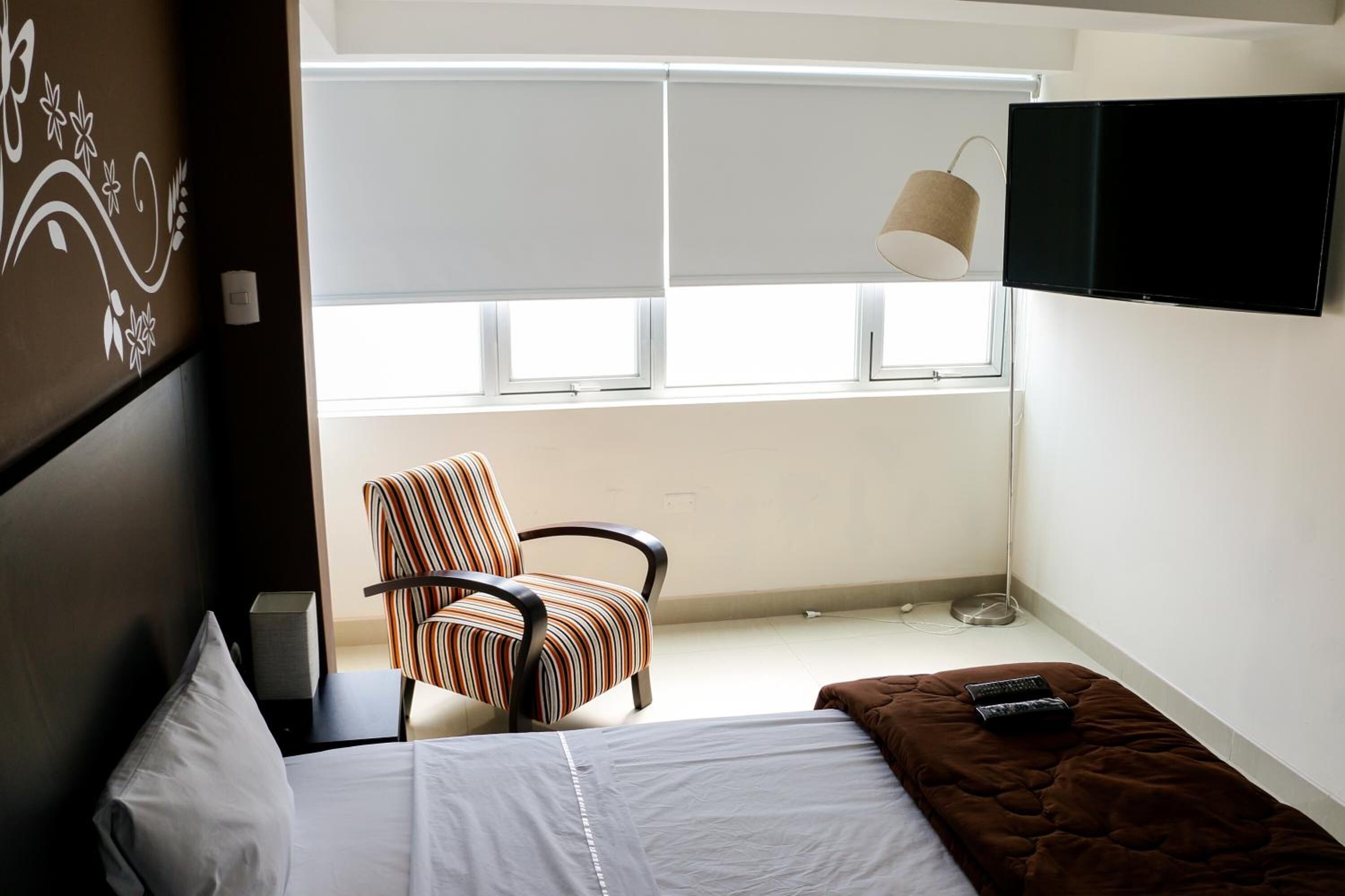 Habitación de hotel con cama, una silla, un televisor y un ventanal con persianas.