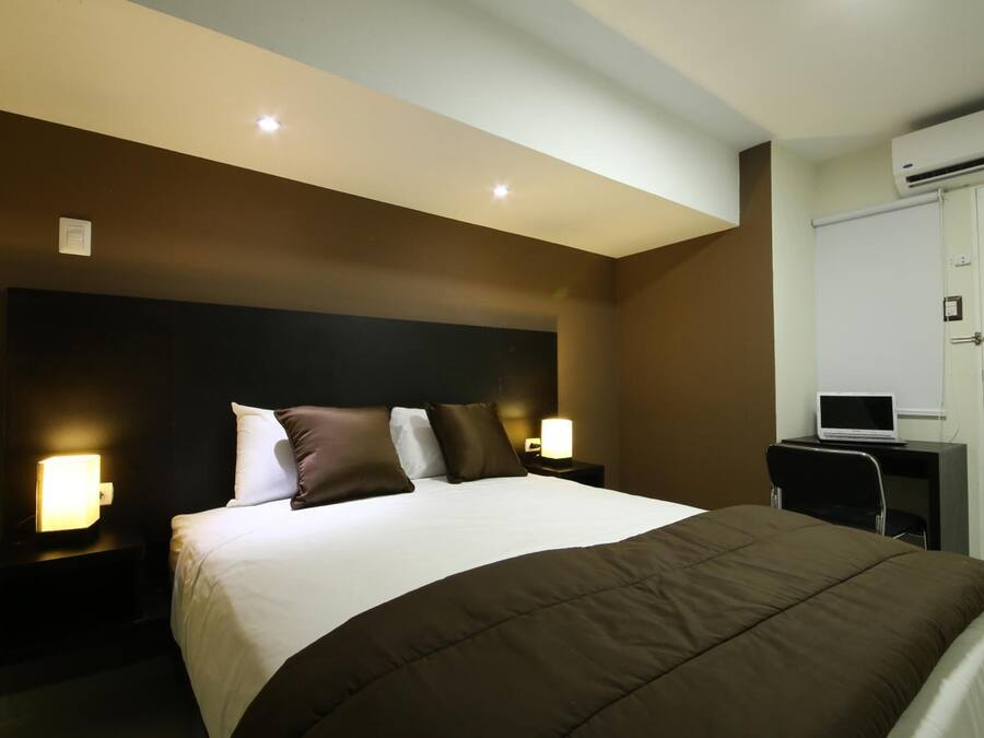A hotel room with a large bed, bedside lamps, a desk with a laptop, and an air conditioning unit.