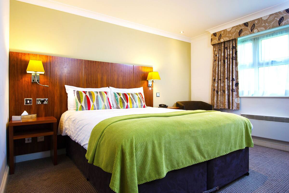 A hotel room with a wooden headboard, two bedside lamps, a bed with colorful pillows, a green blanket, a nightstand, and a window with curtains.