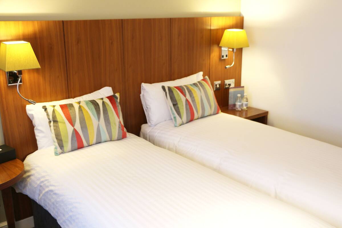 A hotel room with two single beds, each with a white duvet and striped pillows. There are two bedside tables with lamps and a small nightstand with a lamp and a bottle.