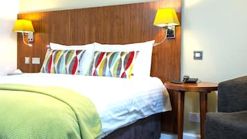 A hotel room with a wooden headboard, a bed with white bedding, a colorful striped pillow, two bedside lamps, a small wooden table with a phone, and a grey armchair.