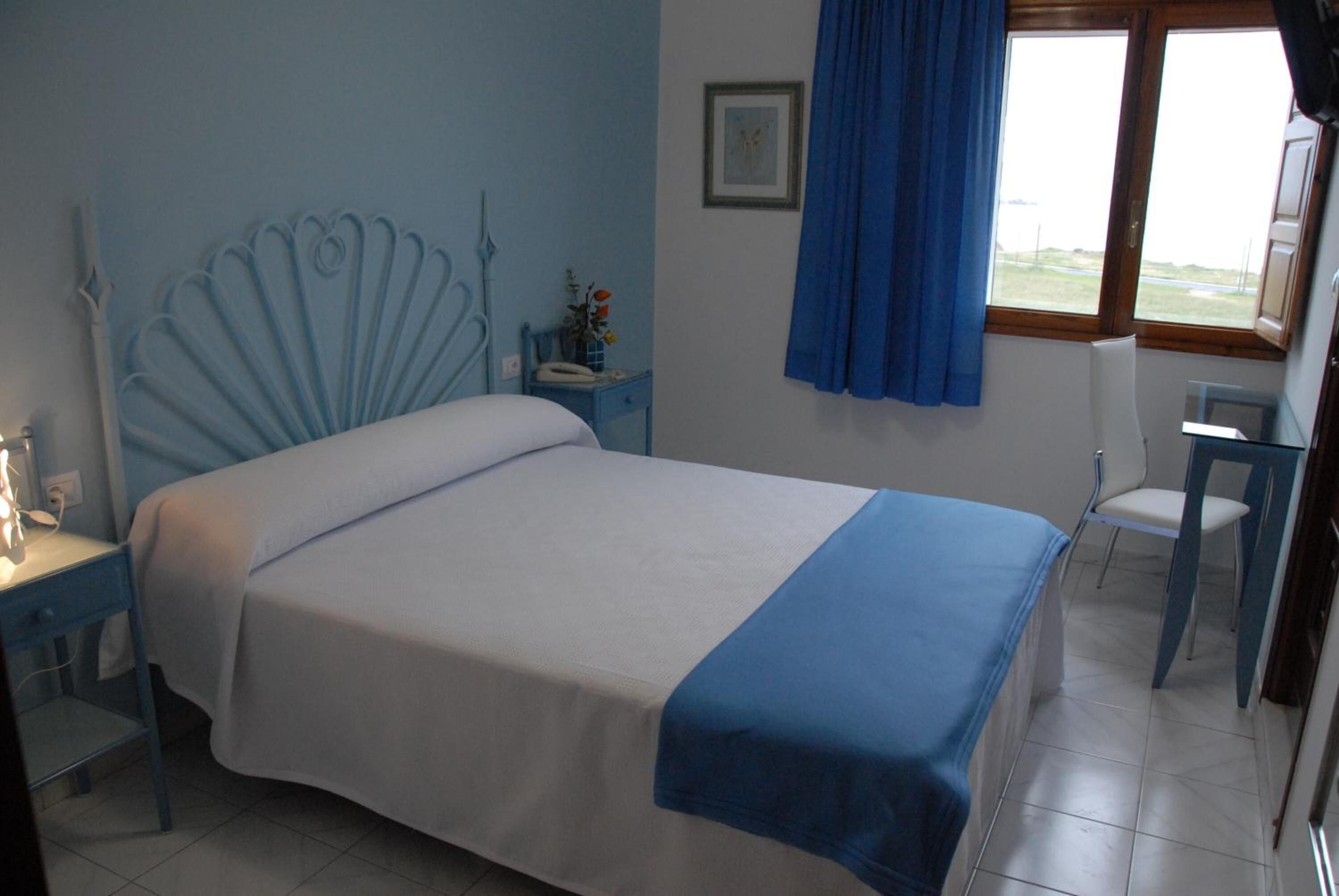 Un dormitorio con una cama, una mesita de noche, una silla, una ventana con cortinas azules y un cuadro en la pared.