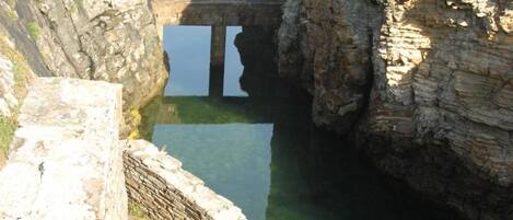 Una estructura de piedra junto a la costa, con un estrecho paso que conduce a una masa de agua, rodeada de acantilados rocosos.