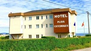 Un hotel llamado "Playa Catedrales" con un diseño moderno, que cuenta con múltiples ventanas y una señalización destacada.