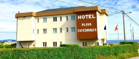 Un hotel llamado "Playa Catedrales" con un diseño moderno, que cuenta con múltiples ventanas y una señalización destacada.