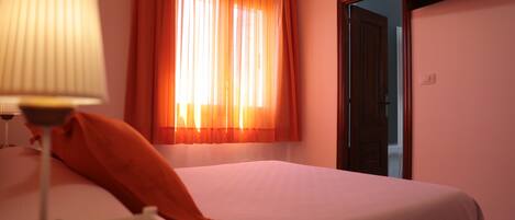 A hotel room with a bed, a lamp, a window with curtains, and a television mounted on the wall.