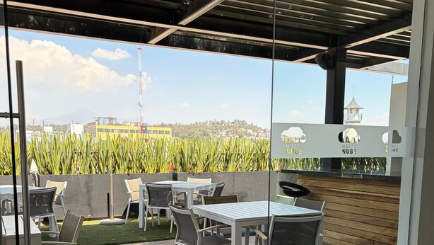 A modern outdoor patio with wooden flooring, white tables, and chairs, overlooking a cornfield and distant buildings. - Hotel Nube, Puebla Centro (Puebla)