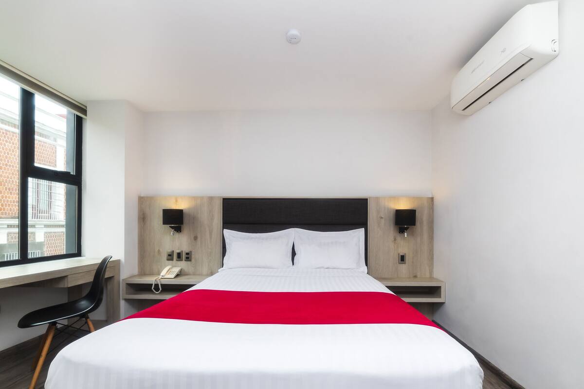 A modern hotel room with a large bed, bedside tables, a desk, and an air conditioning unit.