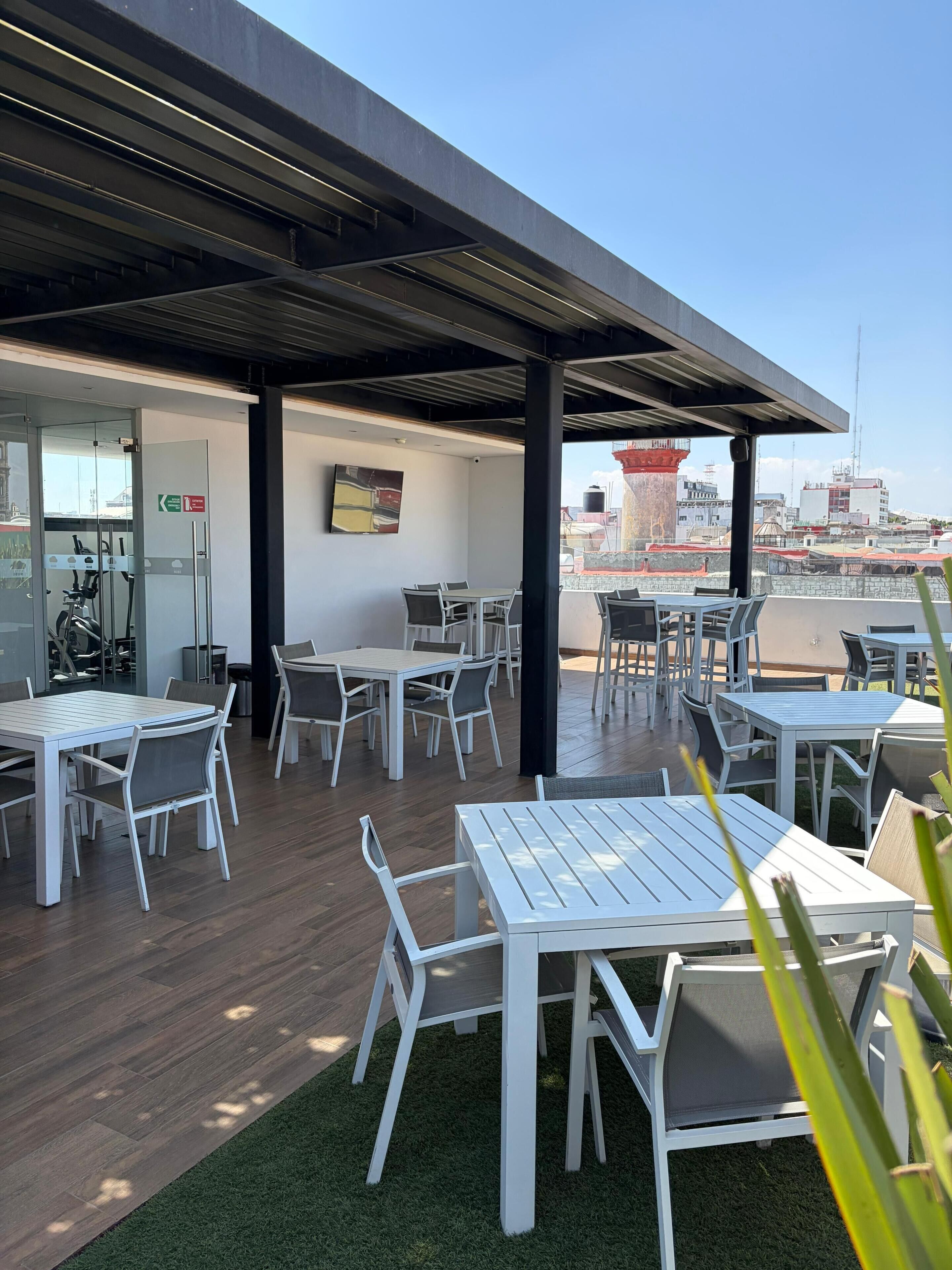 Un patio en la azotea con mesas y sillas, una entrada con puerta de cristal y vistas a edificios al fondo.