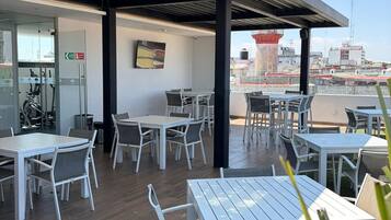 Un patio en la azotea con mesas y sillas, una entrada con puerta de cristal y vistas a edificios al fondo.