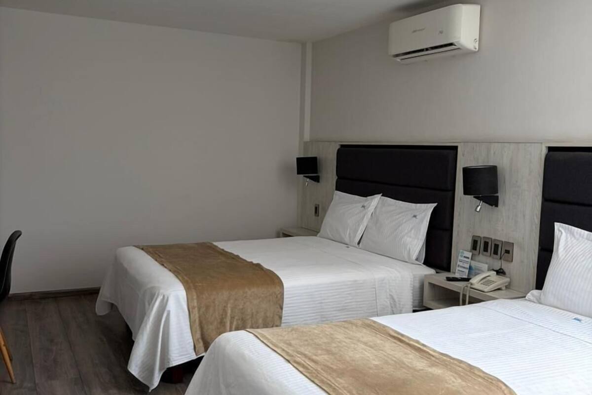 A hotel room with two beds, a wooden floor, a wall-mounted air conditioner, bedside lamps, and a small table with a phone.