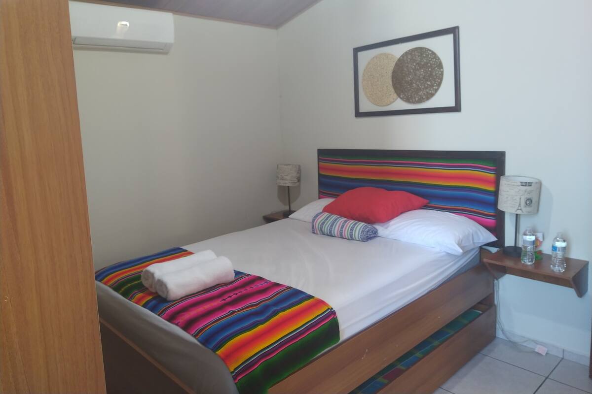 Ein Bett mit einer bunten, gestreiften Decke, einem roten Kissen und einem weißen Handtuch. Ein Nachttisch mit einer Lampe und einer Wasserflasche steht daneben. Der Raum hat weiße Wände und eine Decke mit Holzbalken.