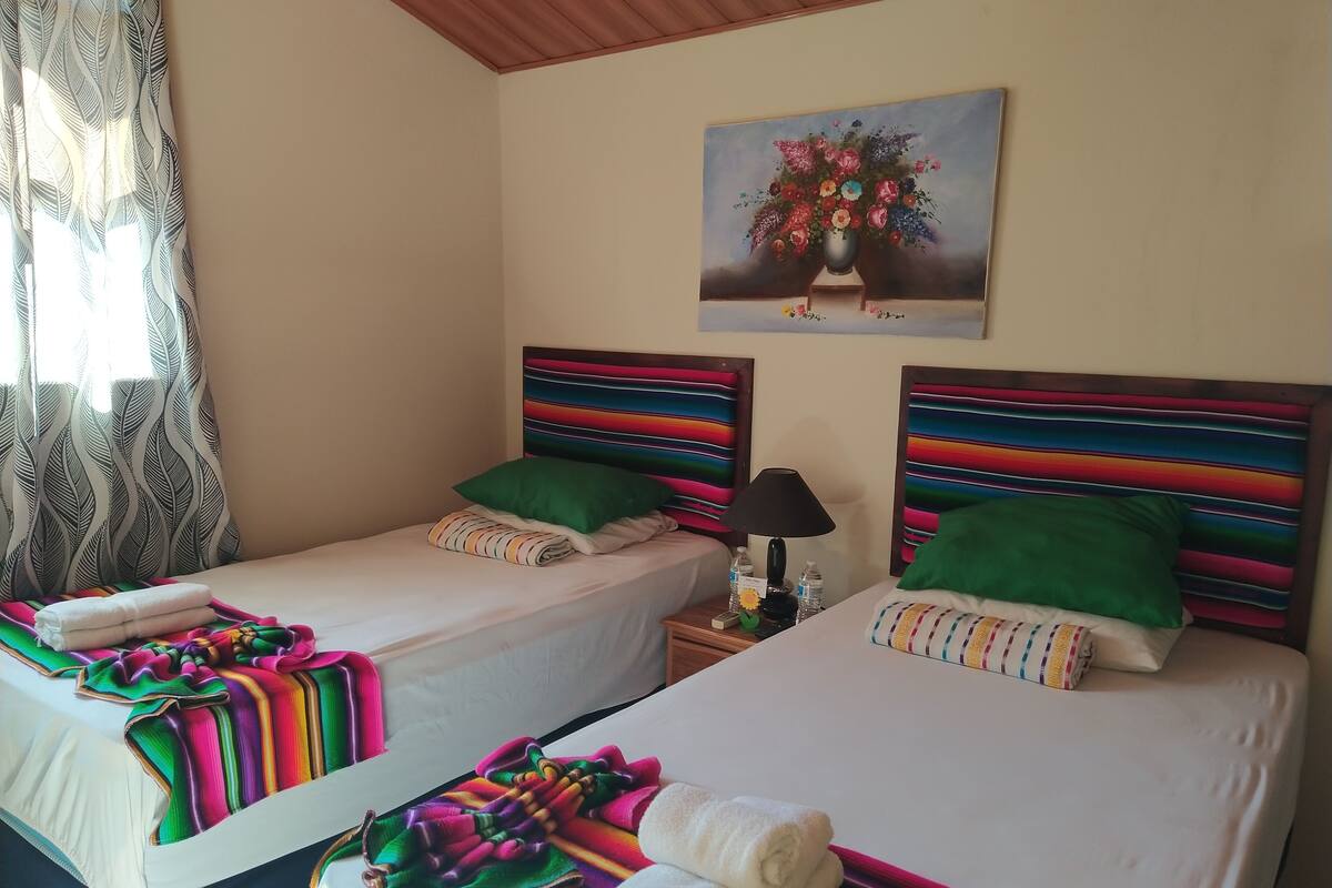 Ein Hotelzimmer mit zwei Betten, jeweils mit farbenfrohen, gestreiften Kopfteilen und Kissen, einem Blumengemälde an der Wand und einem Fenster mit gemusterten Vorhängen.