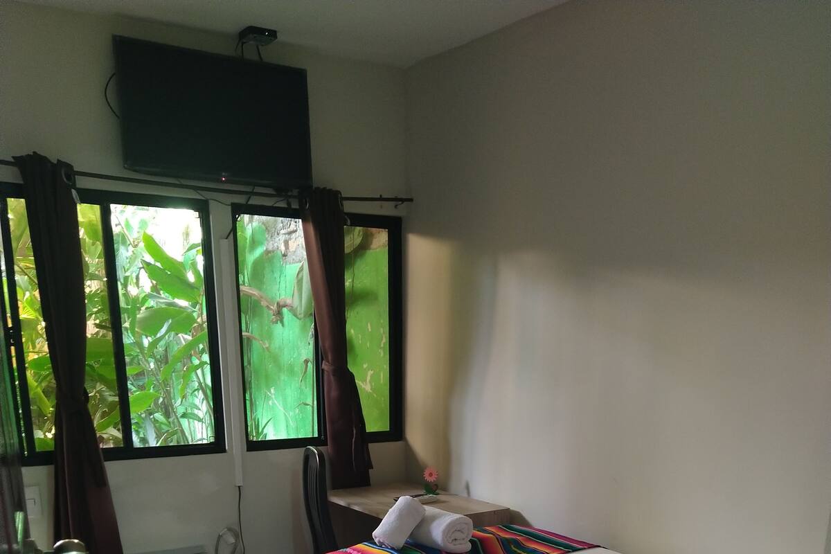 Ein Schlafzimmer mit Bett, Stuhl, einem Fenster mit Vorhängen und einem kleinen Tisch mit einer Blume.