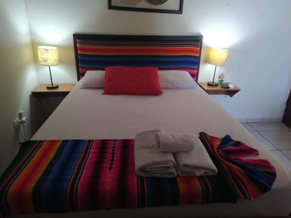 A bed with a colorful striped bedspread, a red pillow, and folded towels on the side. There are two bedside tables with lamps and a small shelf with a phone and a plant.