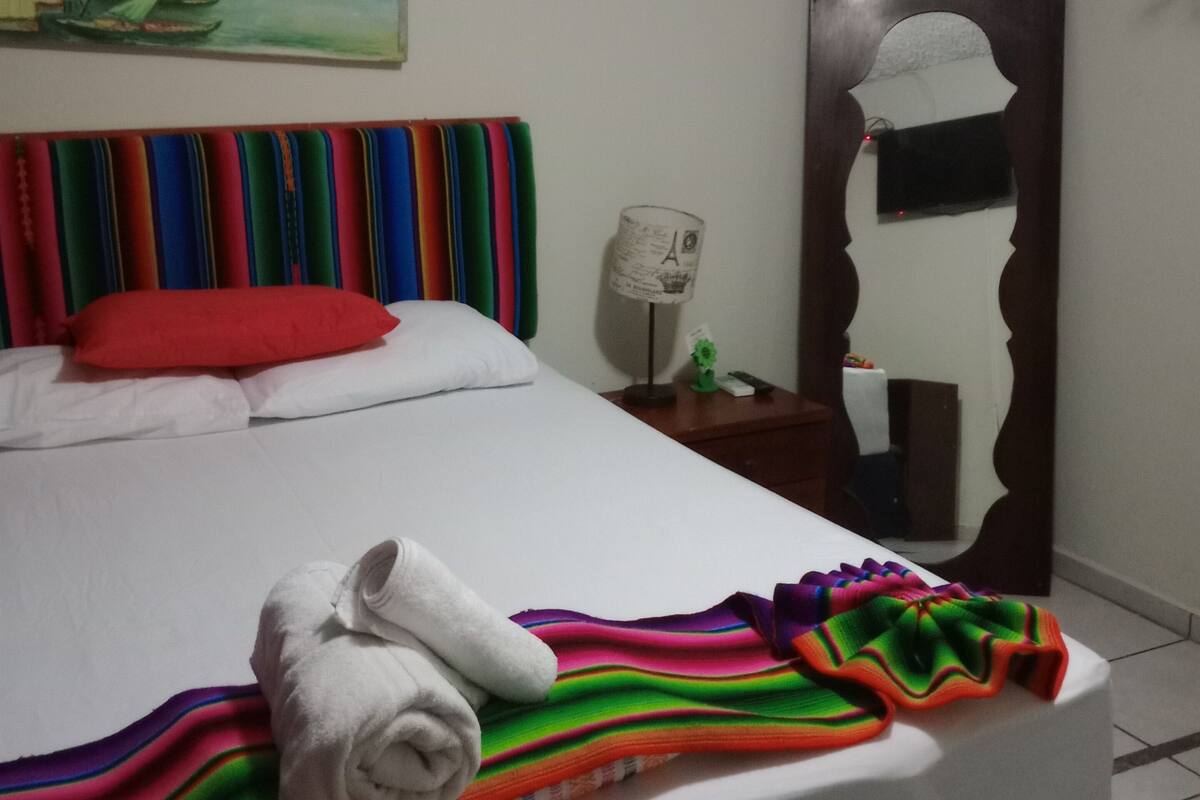 Ein ordentlich bezogenes Bett mit einer farbenfrohen Streifendecke, einem weißen Handtuch, einem roten Kopfkissen und einem Wandbild einer Dorflandschaft.