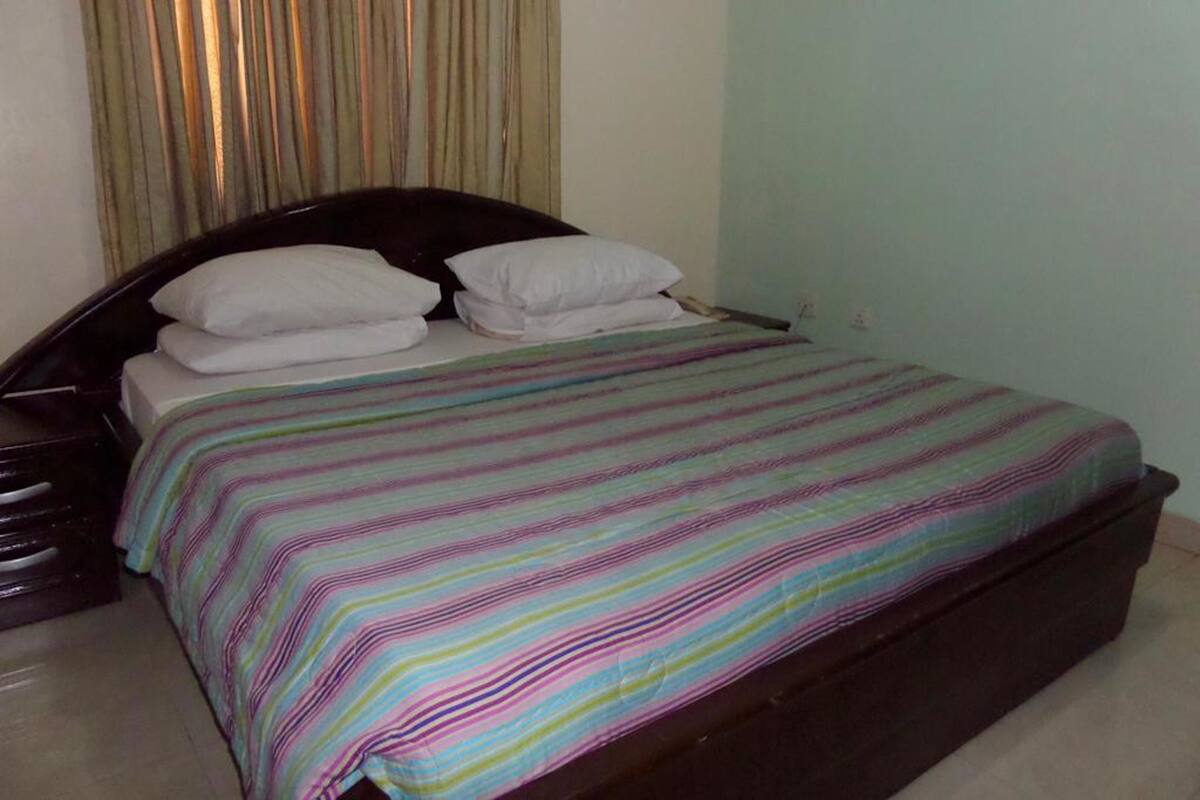A bed with a striped bedspread and two pillows, a dark headboard, and a light-colored wall with a curtain.