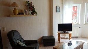 Ein Wohnzimmer mit Ledersessel, Glastisch, Fernseher auf Holzständer, Bücherregal mit Büchern und Pflanze, sowie einem Fenster mit Vorhängen.