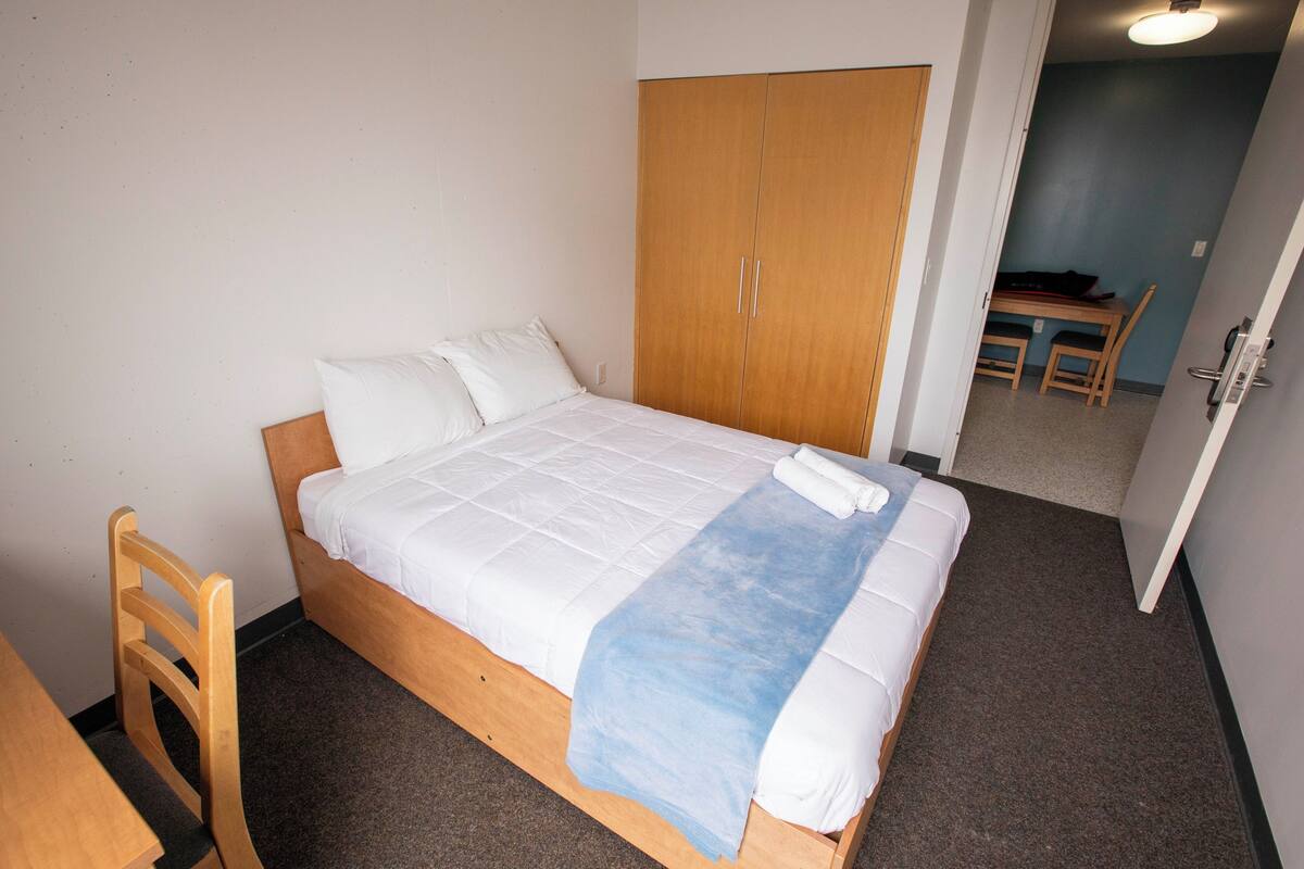 A single bed with white bedding and a blue blanket, a wooden wardrobe, and a small table with chairs in the background.