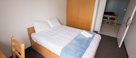 A single bed with white bedding and a blue blanket, a wooden wardrobe, and a small table with chairs in the background.