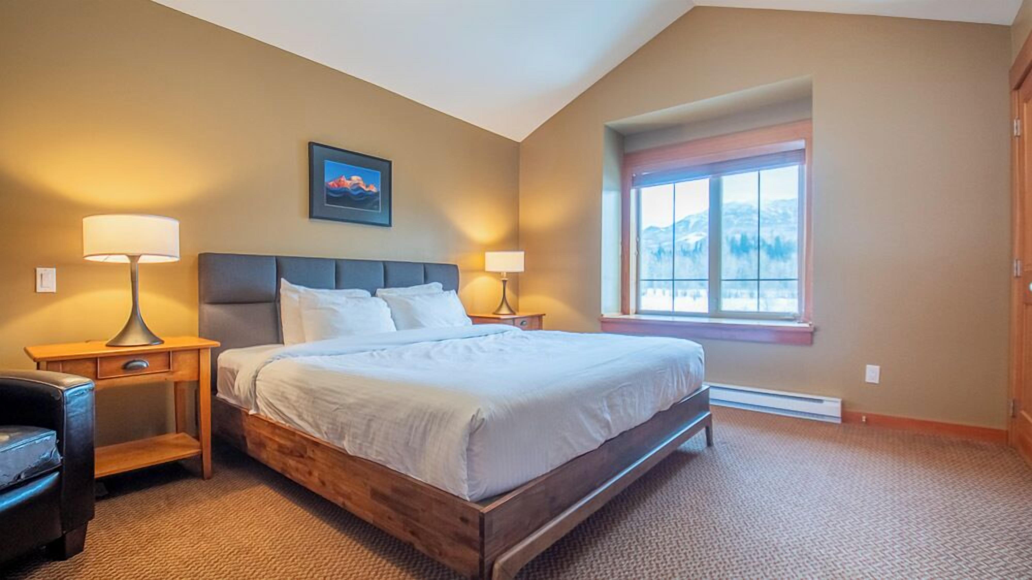 Quarto com cama grande, duas mesas de cabeceira com luminárias, cabeceira de madeira, janela com vista e um quadro na parede.