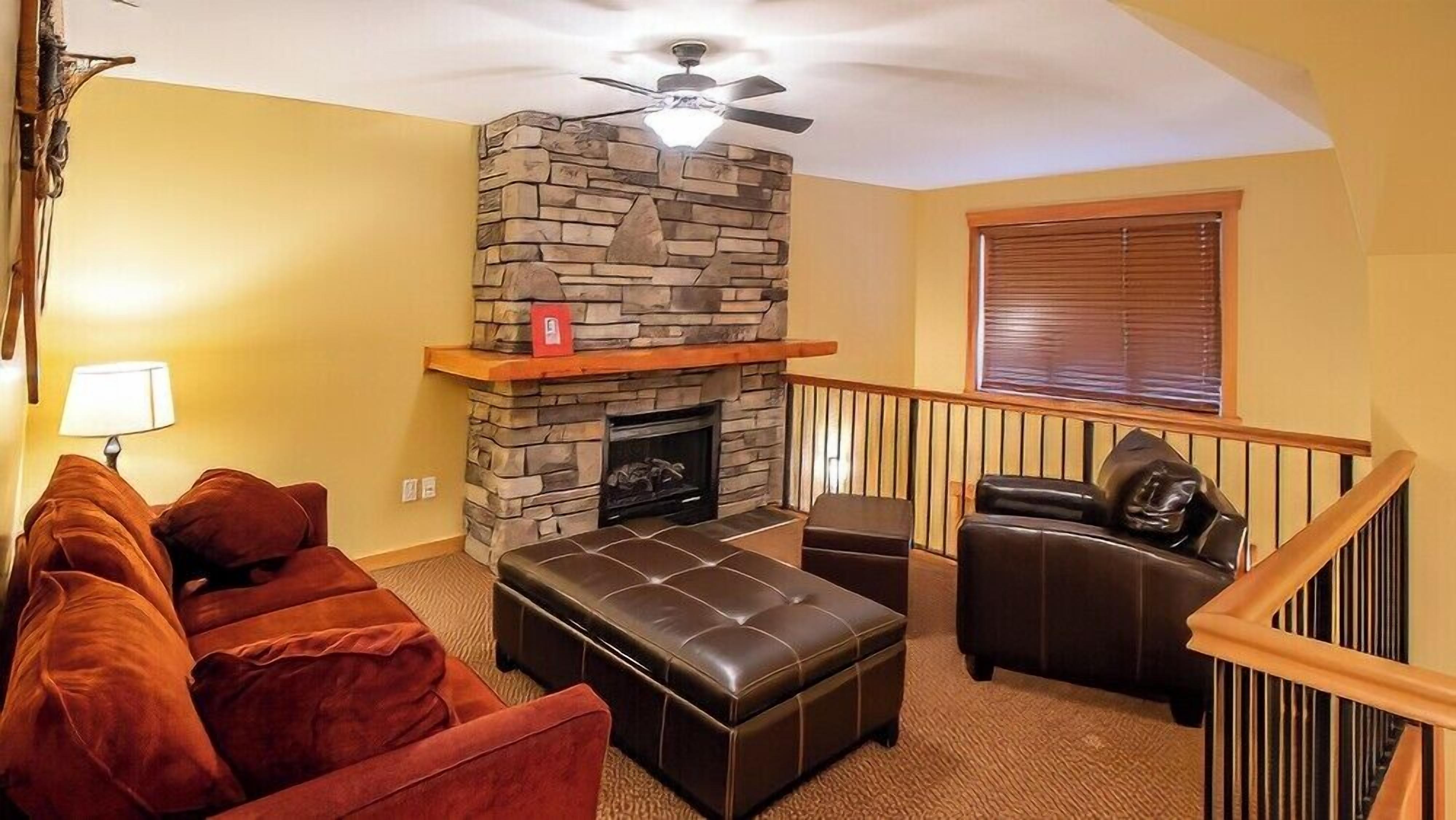 A living room with a stone fireplace, a ceiling fan, a red book on the mantel, a brown leather sofa, and a window with blinds.