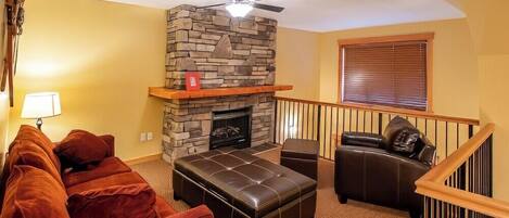 A living room with a stone fireplace, a ceiling fan, a red book on the mantel, a brown leather sofa, and a window with blinds.