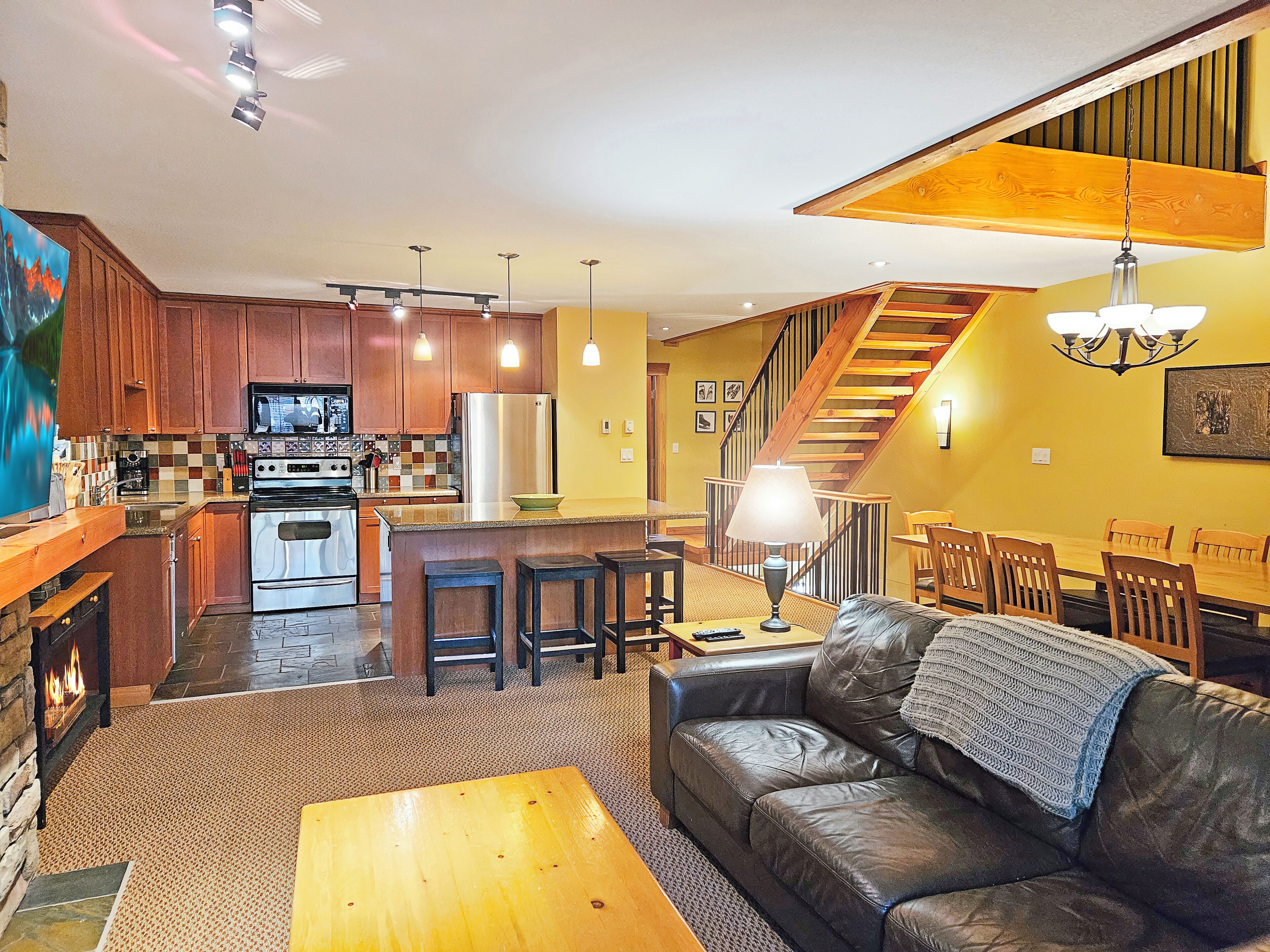 A modern kitchen with wooden cabinets, stainless steel appliances, and pendant lighting. There is a dining area with a leather couch and a staircase leading to another level.