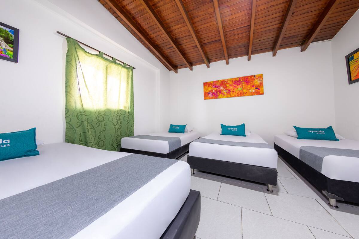 Ein Hotelzimmer mit drei Einzelbetten, jedes mit einem türkisfarbenen Kissen und einer grauen Bettdecke. Das Zimmer verfügt über eine Holzboden, weiße Wände und einen grün gemusterten Vorhang.